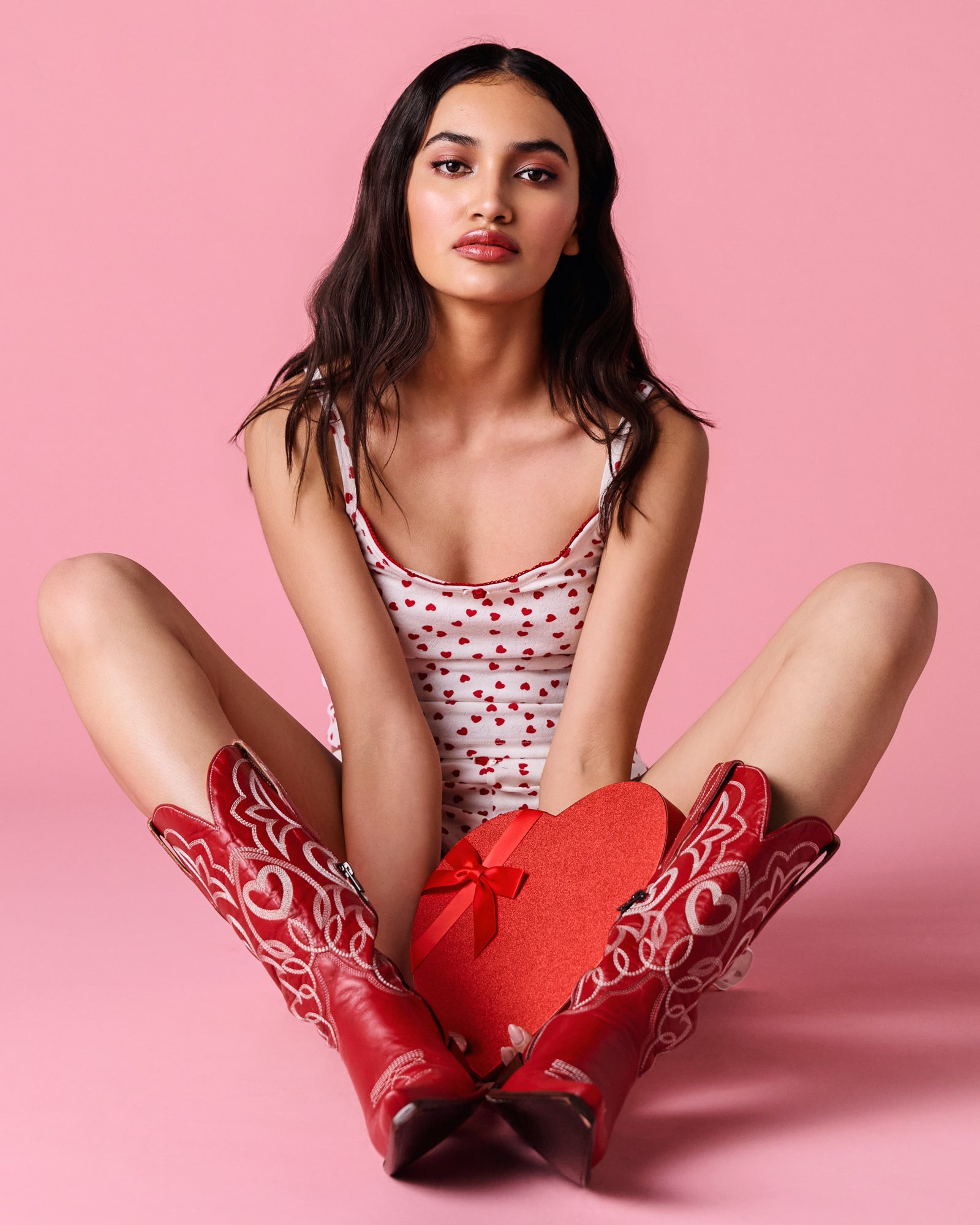 Woman wearing a polka dot pijama and red cowboy boots with a heart-shaped box on a pink background